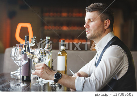 Sommelier sitting at table, looking at wineglass. 137399505
