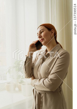 Woman is having a business conversation, standing by the window with her smartphone, feeling thoughtful and socializing while working from home Woman is having a business conversation, standing by the window with her smartphone, feeling thoughtful and socializing while working from home 137399514