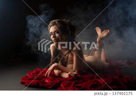 Artistic portrait of a woman in shibari rope tie on red fabric in a dark, smoky studio. 137402175