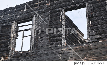 Charred ruins of a burnt wooden log house with empty window frames against an overcast sky. Charred ruins of a burnt wooden log house with empty window frames against an overcast sky. 137404224