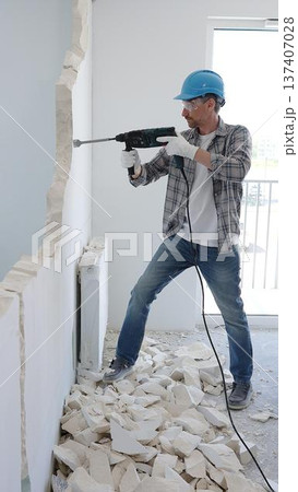 Male construction worker wearing grey checked shirt and blue hardhat is demolishing a wall with a hammer drill, creating construction debris on the surrounding floor surface. Renovation concept 137407028