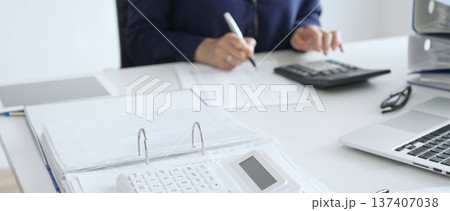 Close up of white calculator over financial documents opposite businesswoman calculating taxes at workplace, binder and glasses on desk. Audit and taxes concept 137407038