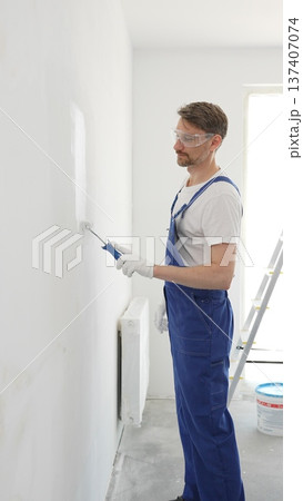 Middle aged man wearing protective white gloves and blue work overalls, applying paint with roller on pristine interior wall during home renovation project, vertical view 137407074