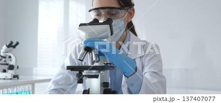 Female scientist in lab coat and safety glasses using microscope, analyzing samples in test tubes in laboratory, conducting scientific research 137407077