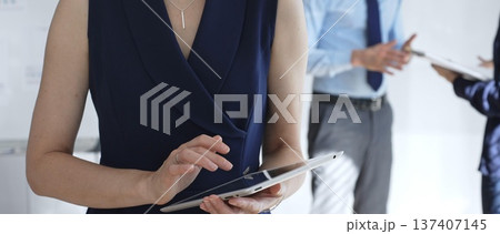Professional businesswoman using a digital tablet in a modern office, interacting with a touchscreen while blurred colleagues collaborate in the background, focusing on tech and teamwork 137407145