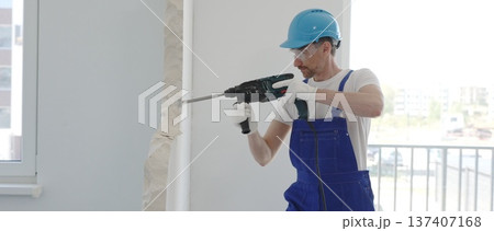 Male construction worker demolishing concrete wall with rotary hammer, wearing work overalls and blue hardhat is generating dust and debris during home renovation work, portrait horizontal view Male construction worker demolishing concrete wall with rotary hammer, wearing work overalls and blue hardhat is generating dust and debris during home renovation work, portrait horizontal view 137407168