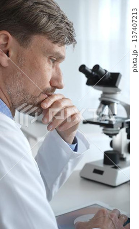 Confident male mature scientist is thinking and analyzing data on a tablet in his laboratory, with a microscope in the background, suggesting advanced scientific research. Medicine and science concept Confident male mature scientist is thinking and analyzing data on a tablet in his laboratory, with a microscope in the background, suggesting advanced scientific research. Medicine and science concept 137407213