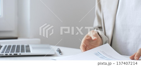 Businesswoman analyzing financial reports, reviewing charts on laptop and paper documents while seated at desk in office workspace. Business people concept Businesswoman analyzing financial reports, reviewing charts on laptop and paper documents while seated at desk in office workspace. Business people concept 137407214