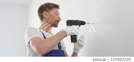 Middle aged male construction worker in a blue jumpsuit, safety goggles, and work gloves is screwing a screw into the wall using a cordless screwdriver, carrying out a careful home renovation with 137407220
