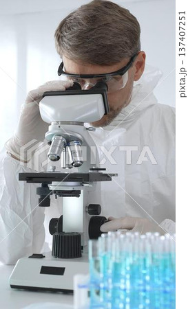 A male medical virologist in a medical gown and gloves examines viral samples under a microscope while conducting research in a sterile laboratory setting A male medical virologist in a medical gown and gloves examines viral samples under a microscope while conducting research in a sterile laboratory setting 137407251