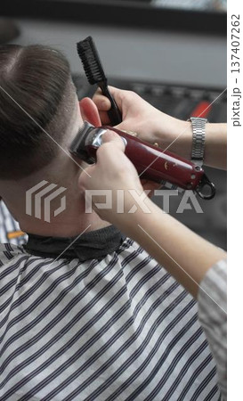 Professional barber grooming client's hair using electric clipper, delivering precise cut with skill in contemporary salon setting, close-up view 137407262