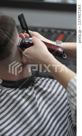 Professional barber grooming client's hair using electric clipper, delivering precise cut with skill in contemporary salon setting, close-up view 137407263