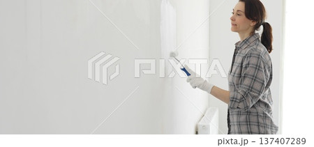 Middle aged happy woman wearing checked shirt and white gloves is painting white wall with paint roller, experiencing home renovation and transforming living space Middle aged happy woman wearing checked shirt and white gloves is painting white wall with paint roller, experiencing home renovation and transforming living space 137407289