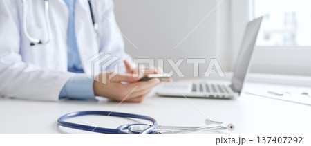 Closeup of blue stethoscope is lying on the table near female doctor checking smartphone representing digital healthcare workflow in well lit clinical workspace. Medicine and health care 137407292