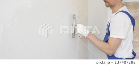 Construction worker plastering white wall, wearing blue overalls and protective white gloves during apartment renovation project Construction worker plastering white wall, wearing blue overalls and protective white gloves during apartment renovation project 137407298