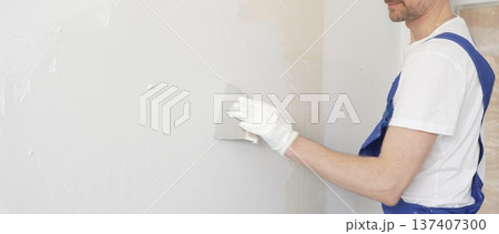 Construction worker plastering white wall, wearing blue overalls and protective white gloves during apartment renovation project 137407300