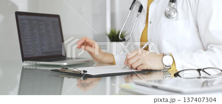 Doctor wearing stethoscope and yellow sweater under white lab coat, making notes on a clipboard in a bright medical office. Medicine, healthcare and science concept 137407304