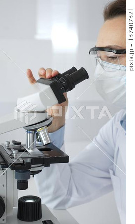 A female scientist in a protective mask and glasses carefully examines a sample under a microscope while conducting research in a modern laboratory. Concept of medicine and science A female scientist in a protective mask and glasses carefully examines a sample under a microscope while conducting research in a modern laboratory. Concept of medicine and science 137407321