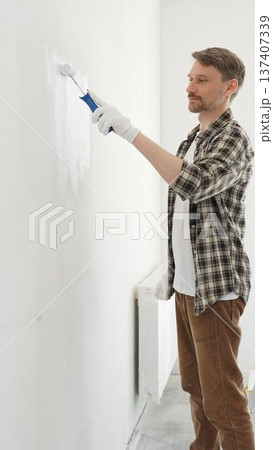 Middle aged man wearing gloves is painting a white wall with a paint roller, working on a home renovation project, creating a fresh and clean look 137407339