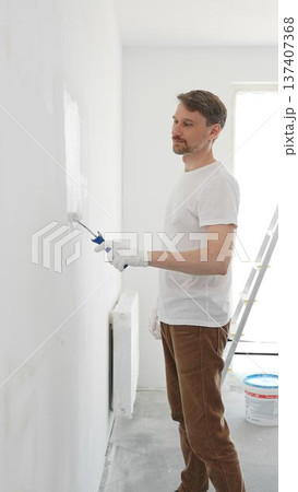 Middle aged man wearing white gloves and t-shirt, is painting white wall with paint roller, experiencing home renovation and transforming living space 137407368
