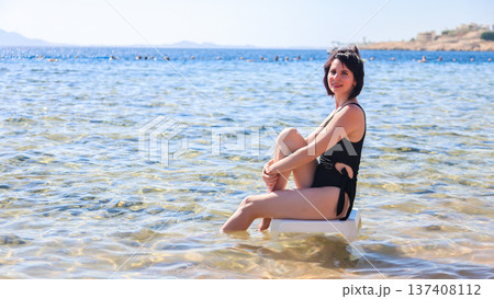 Elegant woman in a black swimsuit sitting on a plastic chair, enjoying the serene sea view, capturing the essence of summer relaxation and beach vibes Elegant woman in a black swimsuit sitting on a plastic chair, enjoying the serene sea view, capturing the essence of summer relaxation and beach vibes 137408112