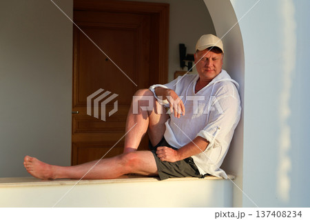 Adult man in a white shirt and cap sitting on a wide windowsill, enjoying the beautiful sunset, reflecting on life and tranquility in the golden hour Adult man in a white shirt and cap sitting on a wide windowsill, enjoying the beautiful sunset, reflecting on life and tranquility in the golden hour 137408234