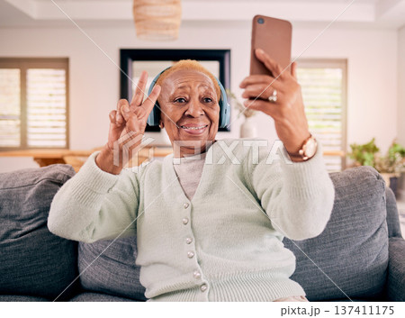 Headphones, selfie and senior woman with peace sign in home listening to music, audio and radio on sofa. Retirement, emoji and happy person in living room take photo for social media and online post 137411175