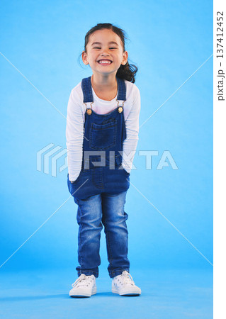 Excited girl child, fashion and studio portrait with jeans, dungaree and trendy style by blue background. Female kid model, smile and happy for clothes, aesthetic and youth culture by backdrop 137412452