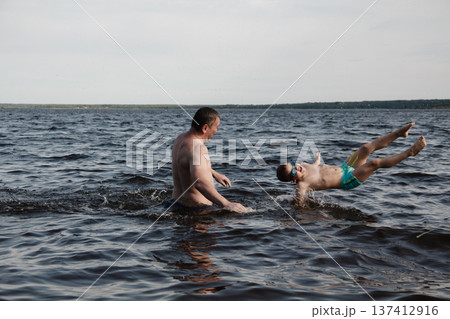 father and son swimming in a lake. man stands calmly in the water nearby. In the background is a green shore, calm waters, and a clear sky. The atmosphere evokes summer relaxation and family warmth. father and son swimming in a lake. man stands calmly in the water nearby. In the background is a green shore, calm waters, and a clear sky. The atmosphere evokes summer relaxation and family warmth. 137412916