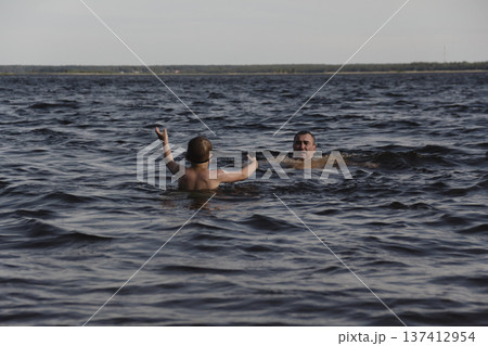father and son swimming in a lake. man stands calmly in the water nearby. In the background is a green shore, calm waters, and a clear sky. The atmosphere evokes summer relaxation and family warmth. father and son swimming in a lake. man stands calmly in the water nearby. In the background is a green shore, calm waters, and a clear sky. The atmosphere evokes summer relaxation and family warmth. 137412954