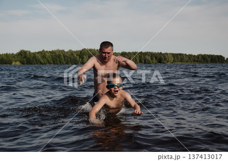 father and son swimming in a lake. man stands calmly in the water nearby. In the background is a green shore, calm waters, and a clear sky. The atmosphere evokes summer relaxation and family warmth. father and son swimming in a lake. man stands calmly in the water nearby. In the background is a green shore, calm waters, and a clear sky. The atmosphere evokes summer relaxation and family warmth. 137413017