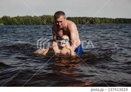 father and son swimming in a lake. man stands calmly in the water nearby. In the background is a green shore, calm waters, and a clear sky. The atmosphere evokes summer relaxation and family warmth. father and son swimming in a lake. man stands calmly in the water nearby. In the background is a green shore, calm waters, and a clear sky. The atmosphere evokes summer relaxation and family warmth. 137413029