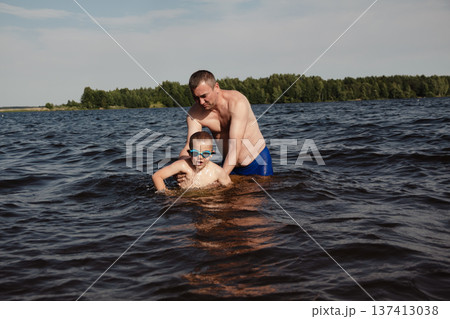 father and son swimming in a lake. man stands calmly in the water nearby. In the background is a green shore, calm waters, and a clear sky. The atmosphere evokes summer relaxation and family warmth. 137413038