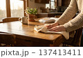 Person wipes down wooden table in kitchen with sunlight streaming through windows. Scene includes houseplant and mug beside open book. Concept of home cleaning, organization, interior design. 137413411