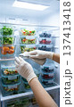 Hands in gloves clean and organize food containers in a fridge. Clear shelves hold salads, berries, and vegetables. Concept of meal prep, healthy eating, food storage solutions. 137413418