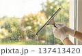 Person cleans window with squeegee in bright light at home. Clear view of outdoor greenery visible through glass. Concept of home maintenance, cleaning services, and window care. 137413420