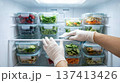 Hand wearing glove arranges clear containers filled with colorful vegetables and fruits in refrigerator. Clean kitchen setting, organized space. Concept of meal prep, health food, cooking. 137413426