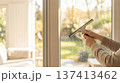 Person uses squeegee to clean glass window in living room with a view of greenery outside. Clear sunlight shines in through window. Concept of cleaning, home improvement, interior design. 137413462