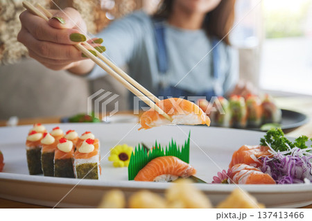 Hands, chopsticks and woman with sushi in restaurant, fine dining and eating in cafe store. Salmon, stick and person on fish, meat and tempura food in plate for lunch, brunch and healthy seafood meal Hands, chopsticks and woman with sushi in restaurant, fine dining and eating in cafe store. Salmon, stick and person on fish, meat and tempura food in plate for lunch, brunch and healthy seafood meal 137413466