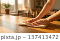 Person wipes wooden table with cloth in cozy room filled with sunlight. Bright setting with soft lighting and furniture in background. Concept of cleaning, home care, interior design. 137413472