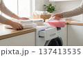 Two people fold clean towels while one pours pink liquid detergent into washing machine. Setting in bright kitchen with natural light and plants. 137413535