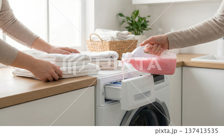 Two people fold clean towels while one pours pink liquid detergent into washing machine. Setting in bright kitchen with natural light and plants. 137413535