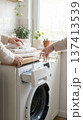 Hands pour liquid detergent into washing machine as towels are stacked on countertop. Bright room filled with plants, windows allowing natural light. 137413539