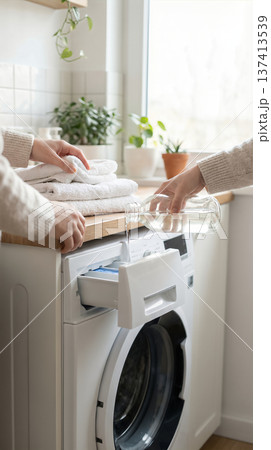 Hands pour liquid detergent into washing machine as towels are stacked on countertop. Bright room filled with plants, windows allowing natural light. 137413539