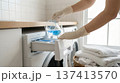 Person wearing gloves pours blue liquid into washing machine drawer in home laundry room. Neat white towels are stacked nearby. Concept of laundry services, cleaning products, household chores. 137413570