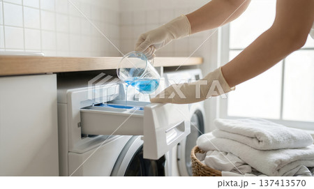 Person wearing gloves pours blue liquid into washing machine drawer in home laundry room. Neat white towels are stacked nearby. Concept of laundry services, cleaning products, household chores. Person wearing gloves pours blue liquid into washing machine drawer in home laundry room. Neat white towels are stacked nearby. Concept of laundry services, cleaning products, household chores. 137413570