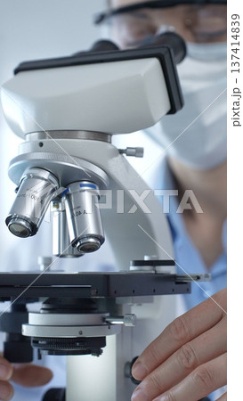 Female scientist wearing lab coat and protective glasses using a modern microscope in a laboratory setting, conducting research and experiments, close up. Health care, medicine and science concept 137414839