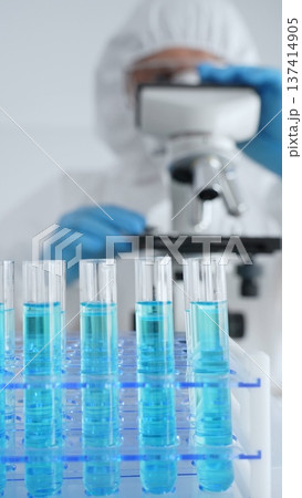 Close up of test tubes with blue liquid opposite a scientific researcher wearing protective gear tuning microscope lenses in laboratory. Medicine, healthcare and science concept Close up of test tubes with blue liquid opposite a scientific researcher wearing protective gear tuning microscope lenses in laboratory. Medicine, healthcare and science concept 137414905