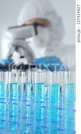 Close up of test tubes with blue liquid opposite a scientific researcher wearing protective gear tuning microscope lenses in laboratory. Medicine, healthcare and science concept 137414927