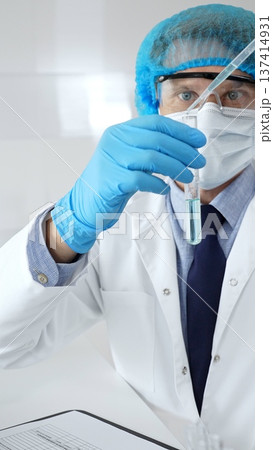 Scientist in protective gear analyzing a test tube of turquoise liquid with a pipette in a modern laboratory setting. Medicine, health care and science concept 137414931
